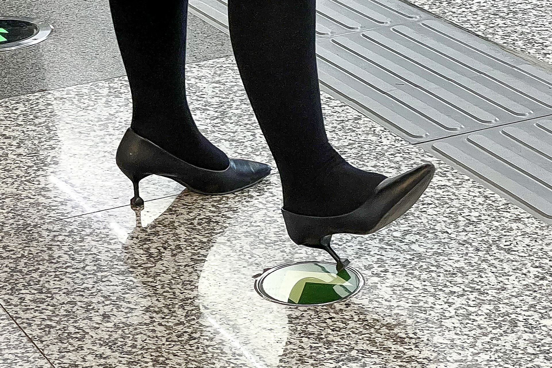 Clearly adult feet in black stockings and black pointed high heels on a reflective floor.