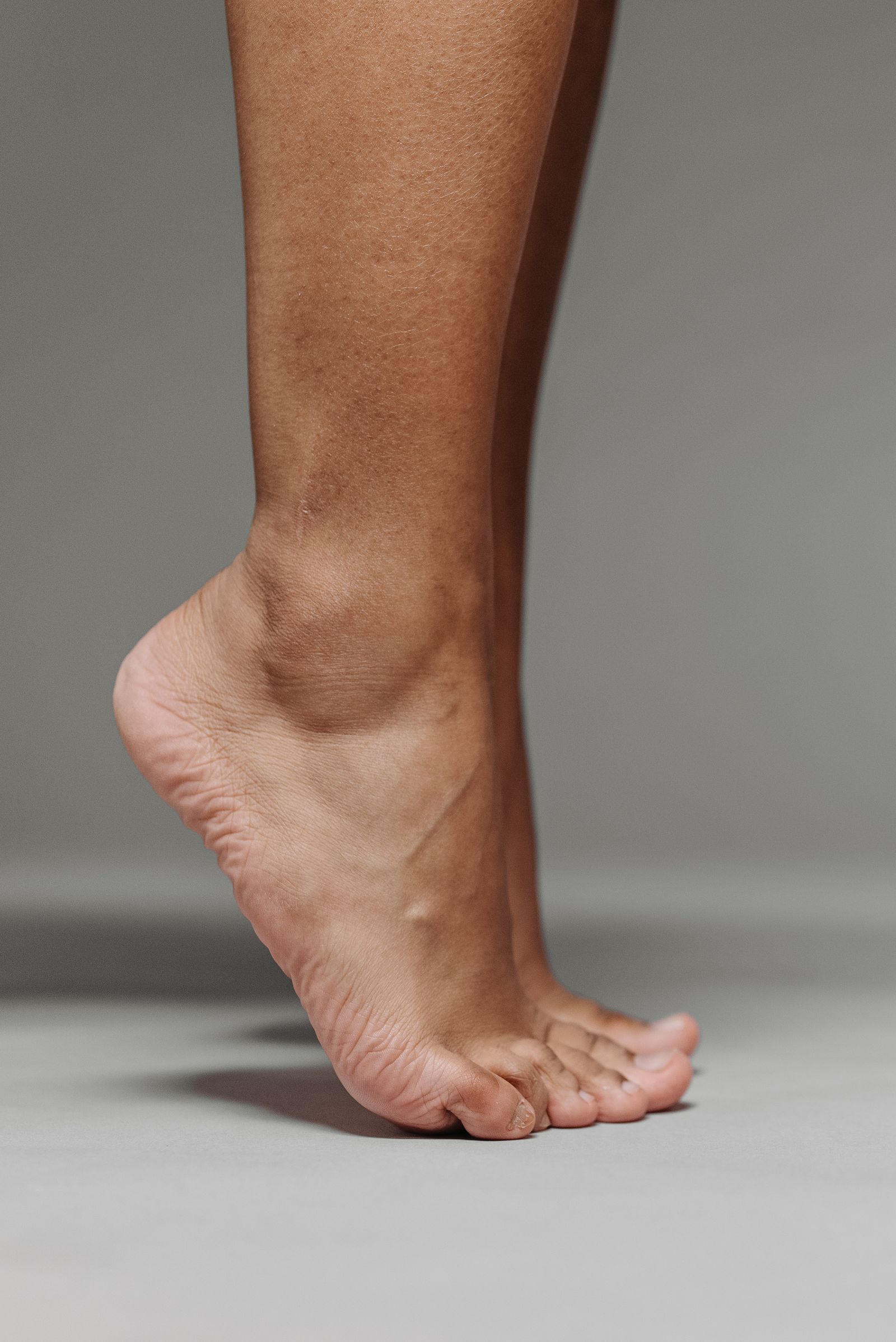 Clearly adult bare foot rising onto the toes in a clean studio close-up with a soft gray background.