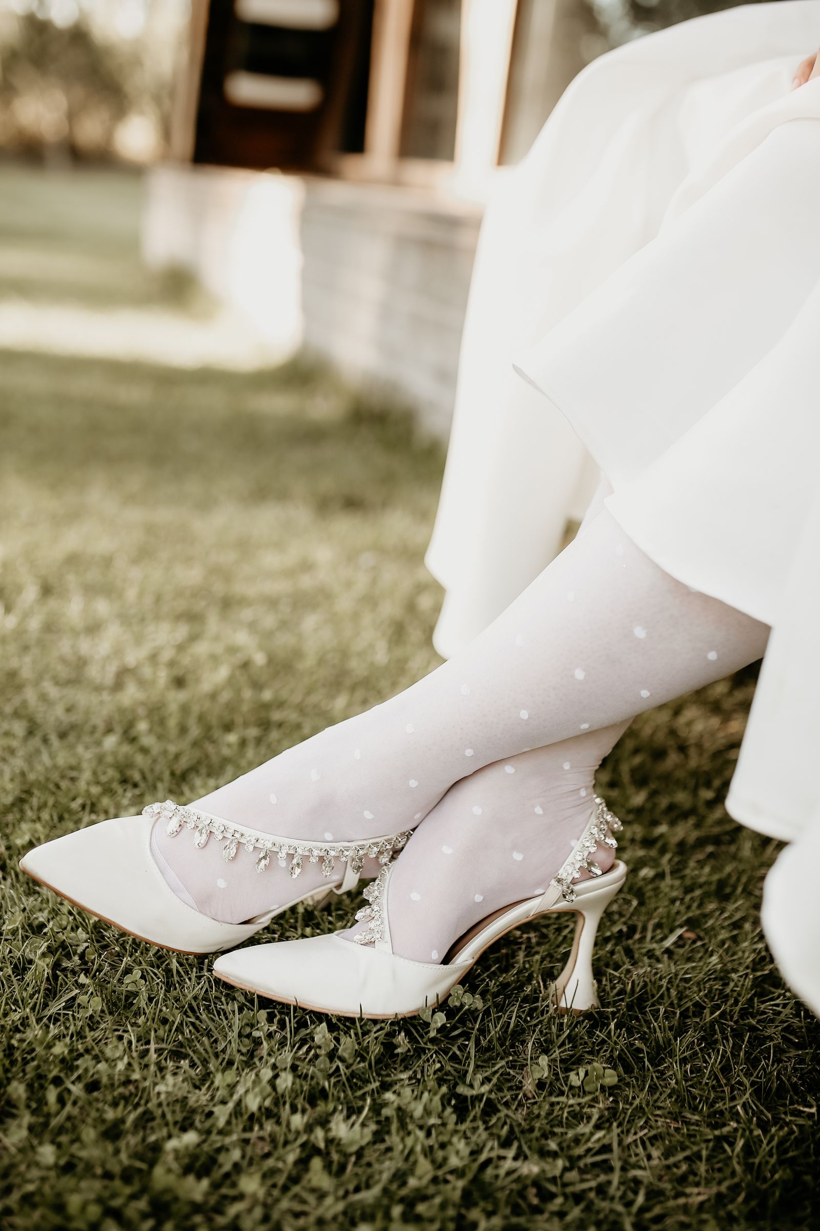 Clearly adult feet in white dotted stockings and embellished white heels in a soft outdoor fashion composition.